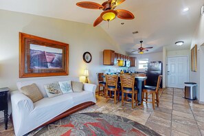 A living room with a sofa, a dining area with chairs, a kitchen with cabinets, and a ceiling fan. - Bayview Condo with Shared Pool, Beach Nearby - Snowbirds Welcome (Pensacola Beach)