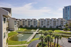 A multi-story residential building with a central courtyard featuring a fountain, surrounded by well-maintained lawns and palm trees. - Bayview Condo with Shared Pool, Beach Nearby - Snowbirds Welcome (Pensacola Beach)