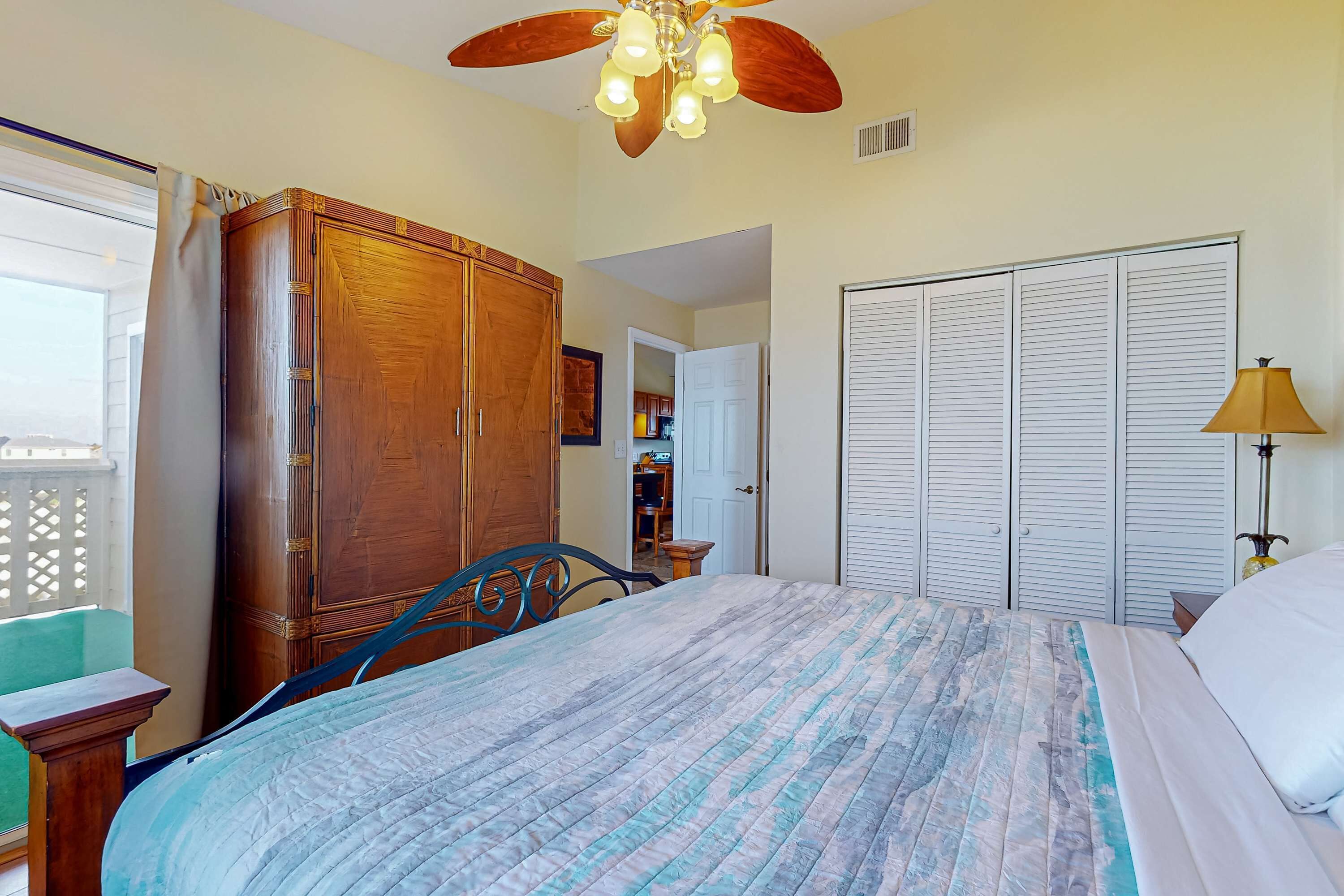 A bedroom with a bed, wooden wardrobe, ceiling fan, and a view of the kitchen through an open door.