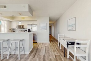 A modern kitchen with white cabinetry, a central island, and bar stools. There are two framed pictures on the wall. - Destin West Bayside Pelican 608 - Gorgeous bayside condo with lazy river and beach access (Fort Walton Beach)