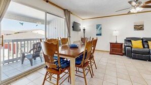 A dining area with a wooden table and chairs, a TV, a ceiling fan, a sofa, and a balcony with chairs. - Cozy Coastal Gulf Condo with Shared Pool and Beach Access (Pensacola Beach)
