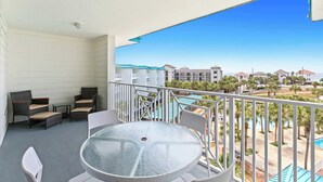 A balcony with wicker chairs, a glass table, and a view of a resort area with buildings and a pool. - Fourth-floor, ocean-view condo with pool, high-speed WiFi, beach access & W&D (Miramar Beach)