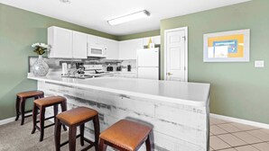 A kitchen with white cabinets, a white island with stools, a microwave, and a refrigerator. - Fourth-floor, ocean-view condo with pool, high-speed WiFi, beach access & W&D (Miramar Beach)
