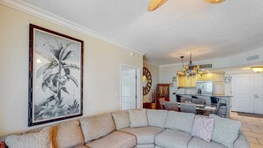 Un salon avec un canapé d’angle, un coin repas avec une table et des chaises, et une cuisine avec des meubles en bois et un lustre.