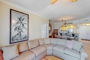 A living room with a sectional sofa, a dining area with a table and chairs, and a kitchen with wooden cabinets and a chandelier. - Gulf-view condo with pool, hot tub, beach access, tennis, balcony, AC, & W/D (Pensacola Beach)