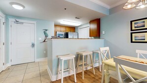 A kitchen with wooden cabinets, a white refrigerator, a microwave, and a sink. There are three white bar stools and a yellow table with chairs. - Beachview Condo with full kitchen, shared pool and beach access (Fort Walton Beach)