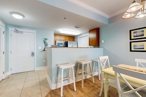 A kitchen with wooden cabinets, a white refrigerator, a microwave, and a sink. There are three white bar stools and a yellow table with chairs. - Beachview Condo with full kitchen, shared pool and beach access (Fort Walton Beach)