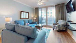 A modern living room with a grey sofa, a patterned armchair, a flat-screen TV mounted on the wall, and a ceiling fan. - Beachview Condo with full kitchen, shared pool and beach access (Fort Walton Beach)