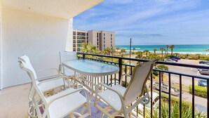 Ein Balkon mit Tisch und Stühlen, der Blick auf einen Strand mit Palmen und ein Gebäude im Hintergrund bietet.
