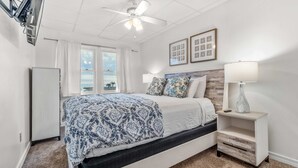 Une chambre à coucher avec un lit, une table de chevet, un ventilateur de plafond, une fenêtre avec des rideaux et des tableaux encadrés accrochés au mur.