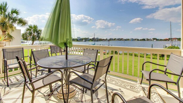 Une terrasse avec une table ronde et des chaises, un parasol vert, et une vue sur une étendue d’eau avec des bâtiments en arrière-plan.