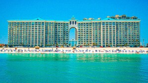 Un gran hotel con varios pisos y un arco central, situado en una playa con numerosas sombrillas y gente.