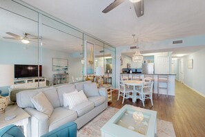 Ein geräumiges Wohnzimmer mit Sofa, Couchtisch und Fernseher. Ein Essbereich mit einer Chandelieber ist vorhanden, durch eine offene Tür ist außerdem eine Küche zu sehen.