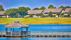 Un quiosco en un muelle sobre un lago, rodeado por un campo de golf y edificios con tejados de paja.