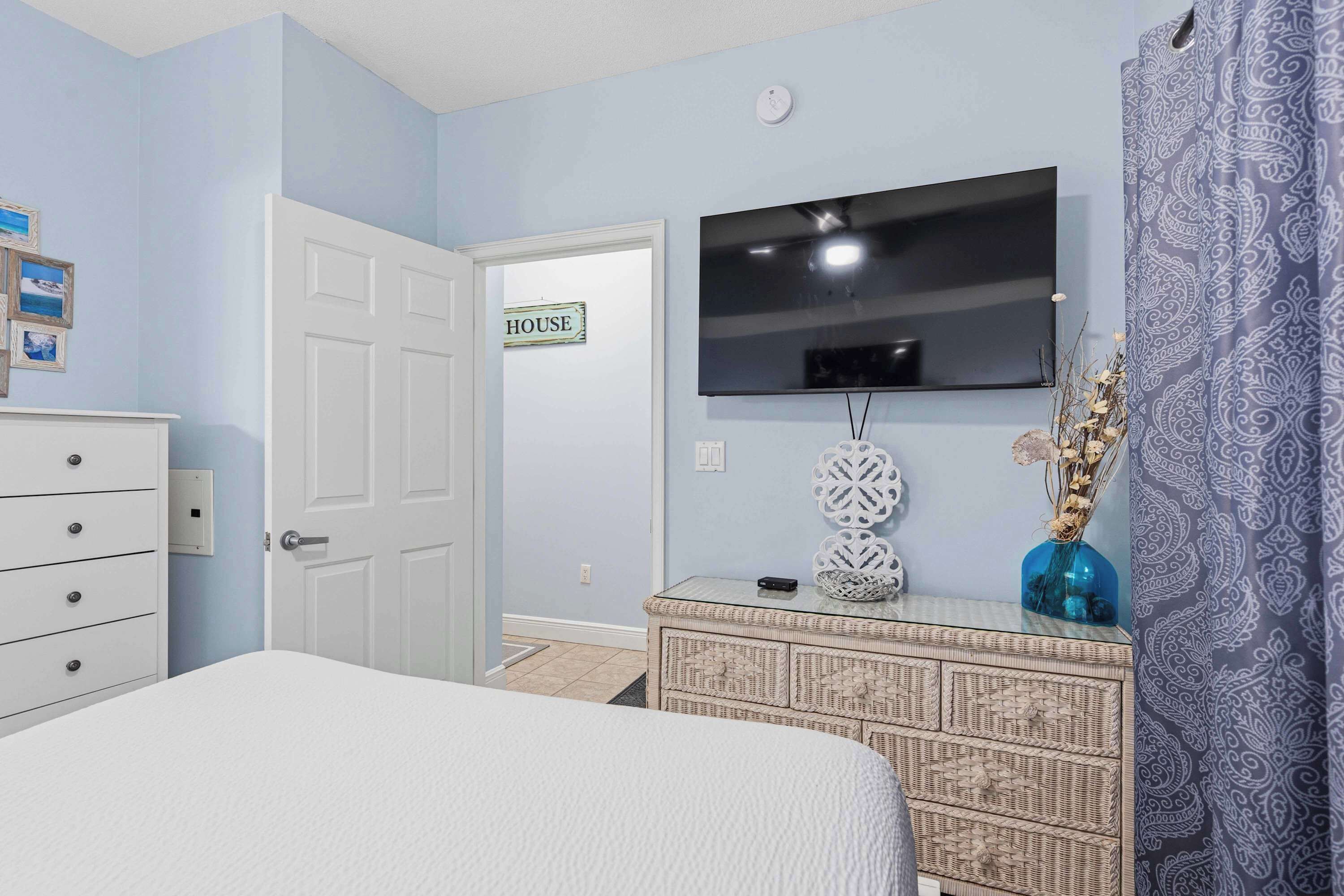 A bedroom with a bed, a dresser, a television on the wall, and a door with a 'HOUSE' sign.
