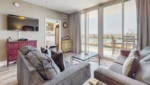 A living room with a sofa, a TV, a ceiling fan, and a view of a pool and beach through large windows. - Gorgeous ground floor beachfront condo, shared pool and beach access (Fort Walton Beach)