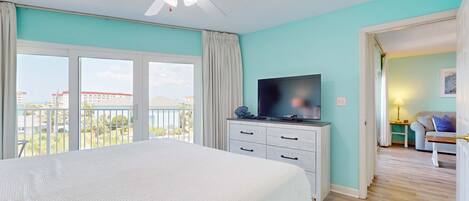 A bedroom with a large bed, a ceiling fan, a TV, and a view of the outdoors.