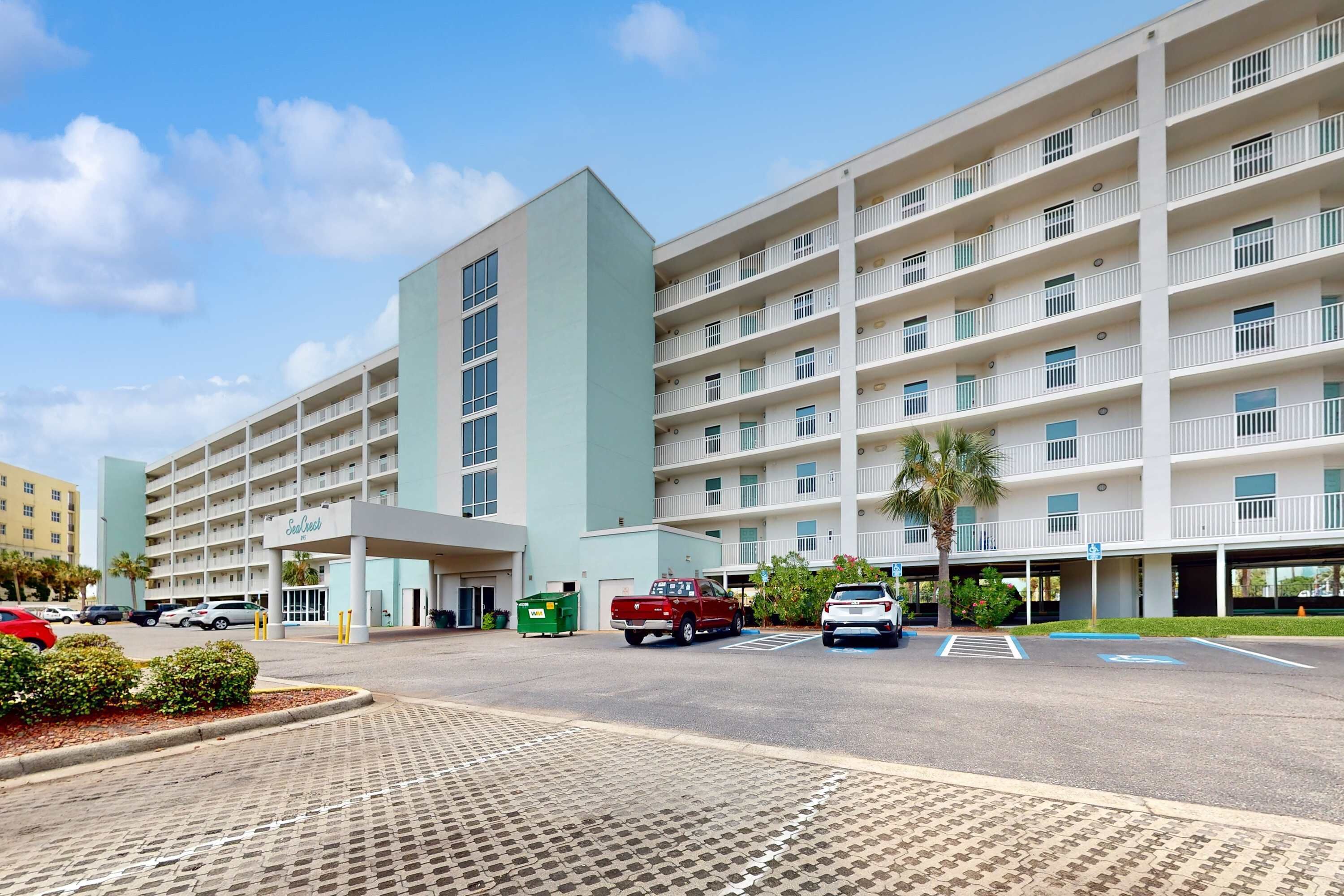 A multi-story hotel with a central entrance, parking area, and a clear sky.