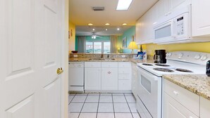 A kitchen with white cabinets, a stove, a microwave, and a window with curtains. - Comfortable Vacation Rental On Okaloosa Island, On The Beach, On-Site Pool (Fort Walton Beach)