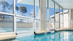 A clear indoor swimming pool with large windows and a view of palm trees. - Comfortable Vacation Rental On Okaloosa Island, On The Beach, On-Site Pool (Fort Walton Beach)