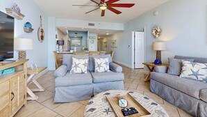 A spacious living room with a sectional sofa, a coffee table, and a ceiling fan. A kitchen area is visible in the background. - Comfortable and cozy beachfront condo w shared amenities (Fort Walton Beach)