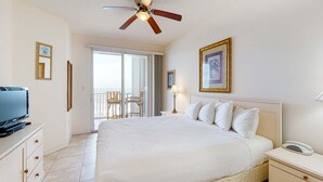 Een hotelkamer met een groot bed, een plafondventilator, een televisie, een nachtkastje met een lamp en een schuifdeur naar een balkon met stoelen.