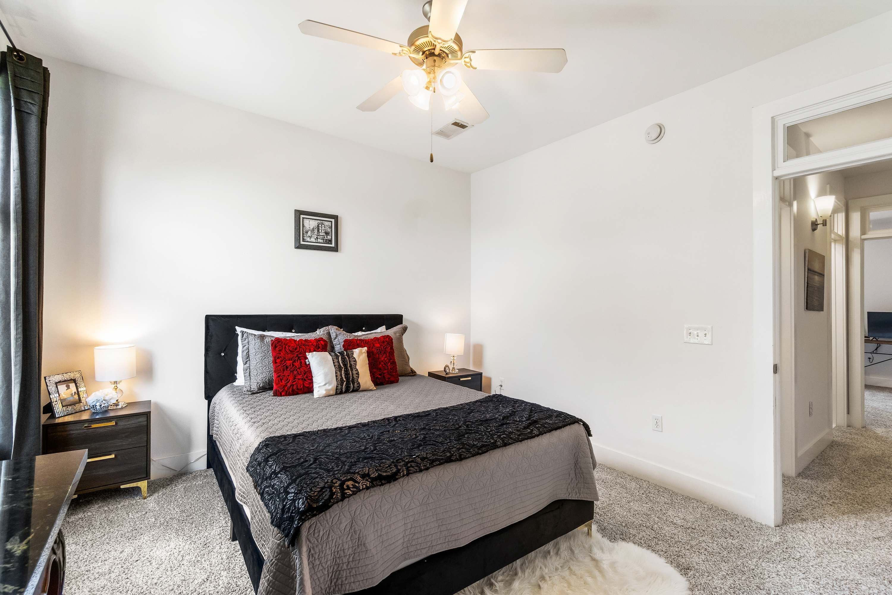 A bedroom with a bed, bedside tables, a ceiling fan, and a door leading to another room.