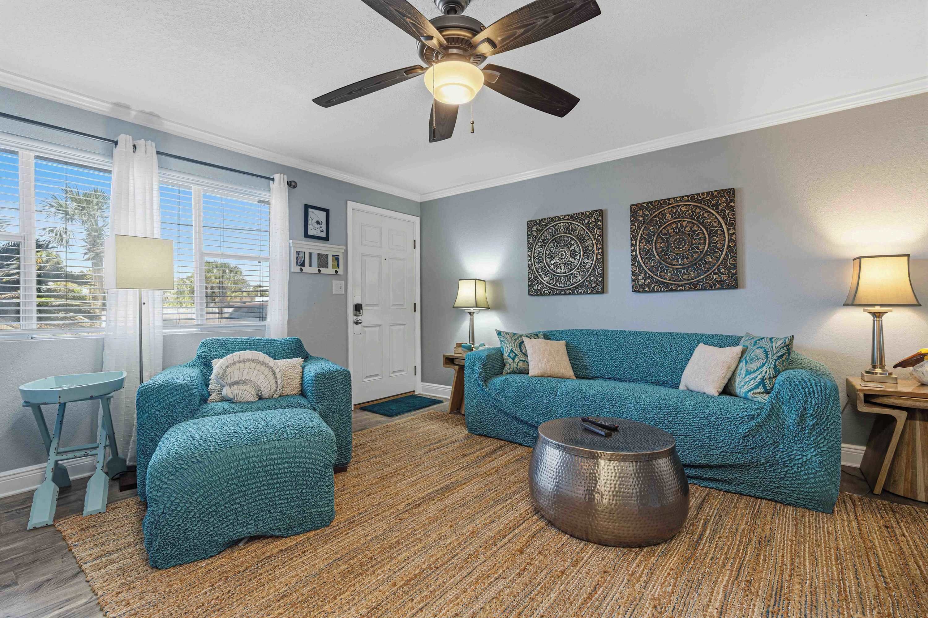 Un salon avec deux canapés de couleur turquoise, une table basse ronde, un ventilateur de plafond et une grande fenêtre avec des rideaux.