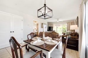 A dining area with a wooden table set for four, a chandelier, and a view of the living room. - Beautiful condo with easy beach access, pool, tennis, courtyard view, & W&D (Miramar Beach)