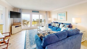 A living room with a blue sofa, a flat-screen TV, a dining table, and a view of the outdoors. - Beachfront ground-floor condo with beach & Gulf views, outdoor pool & tennis (Miramar Beach)