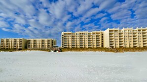 A beachfront hotel with multiple floors and balconies, a sandy beach in the foreground, and a clear sky with clouds. - Beachfront ground-floor condo with beach & Gulf views, outdoor pool & tennis (Miramar Beach)