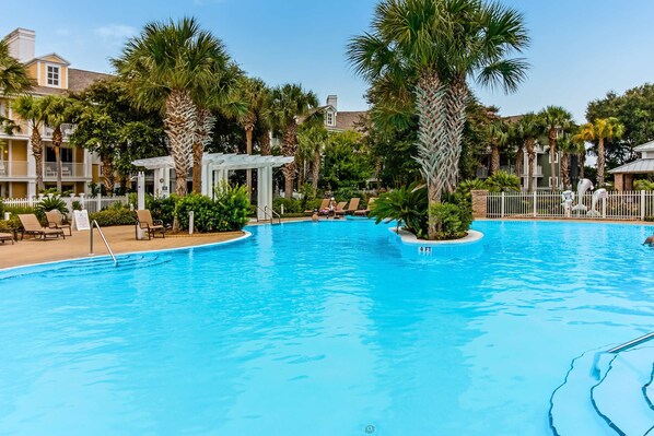 A swimming pool surrounded by palm trees and lounge chairs, with a white fence and a building in the background.