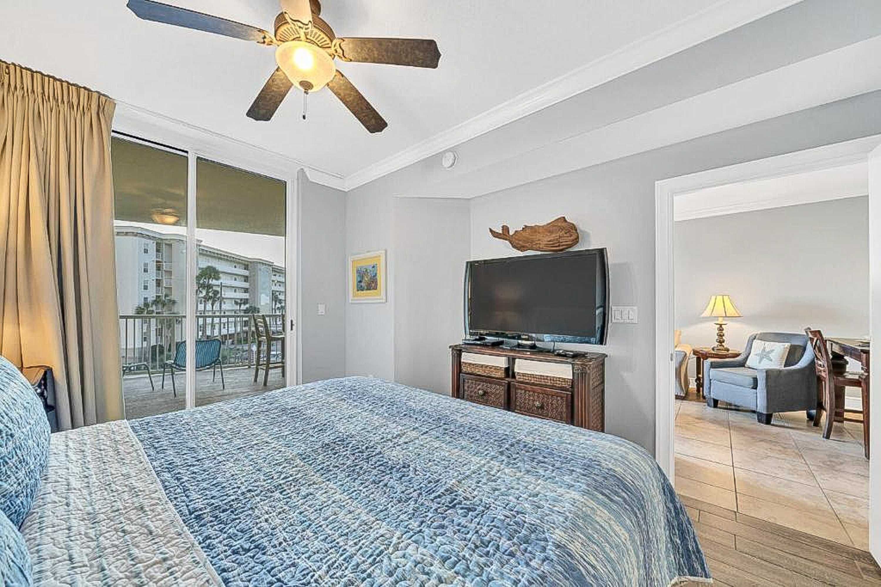 A hotel room with a large bed, a ceiling fan, a television, and a view of the city through the balcony doors.