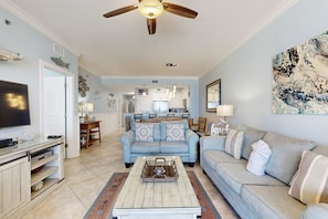 A living room with a TV, a coffee table, and a sofa. There is a kitchen in the background. - gulf front condo steps from the sand with shared pool,hot tub & more (Fort Walton Beach)