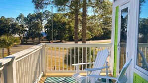 A balcony with white chairs and a green-striped mat, overlooking a street with trees and a stop sign. - Dog-Friendly Florida Beach House Steps from the Water in Carrabelle (Carrabelle)