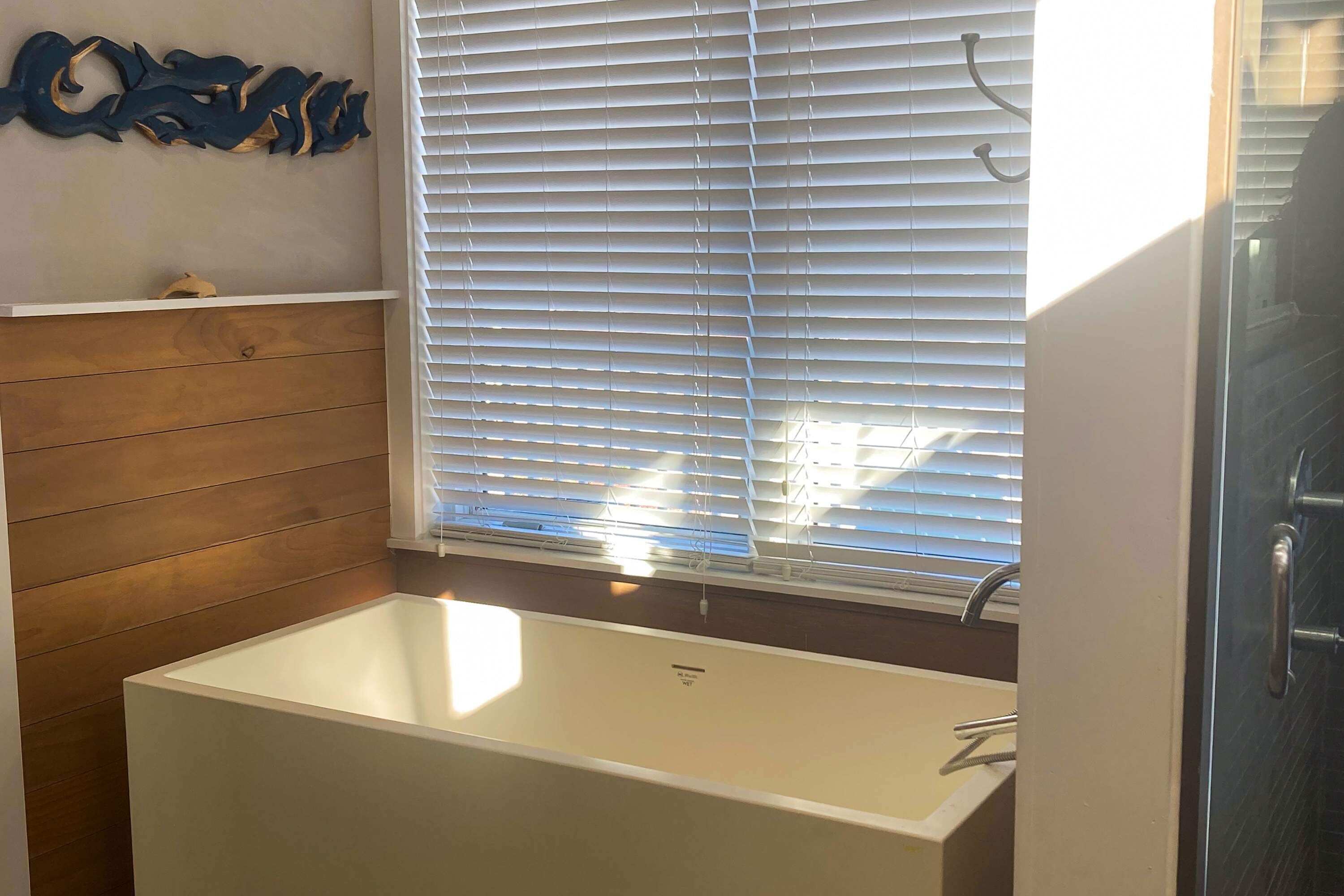 Une salle de bain avec une baignoire blanche, un lambris en bois, un élément décoratif mural et une fenêtre avec des stores.