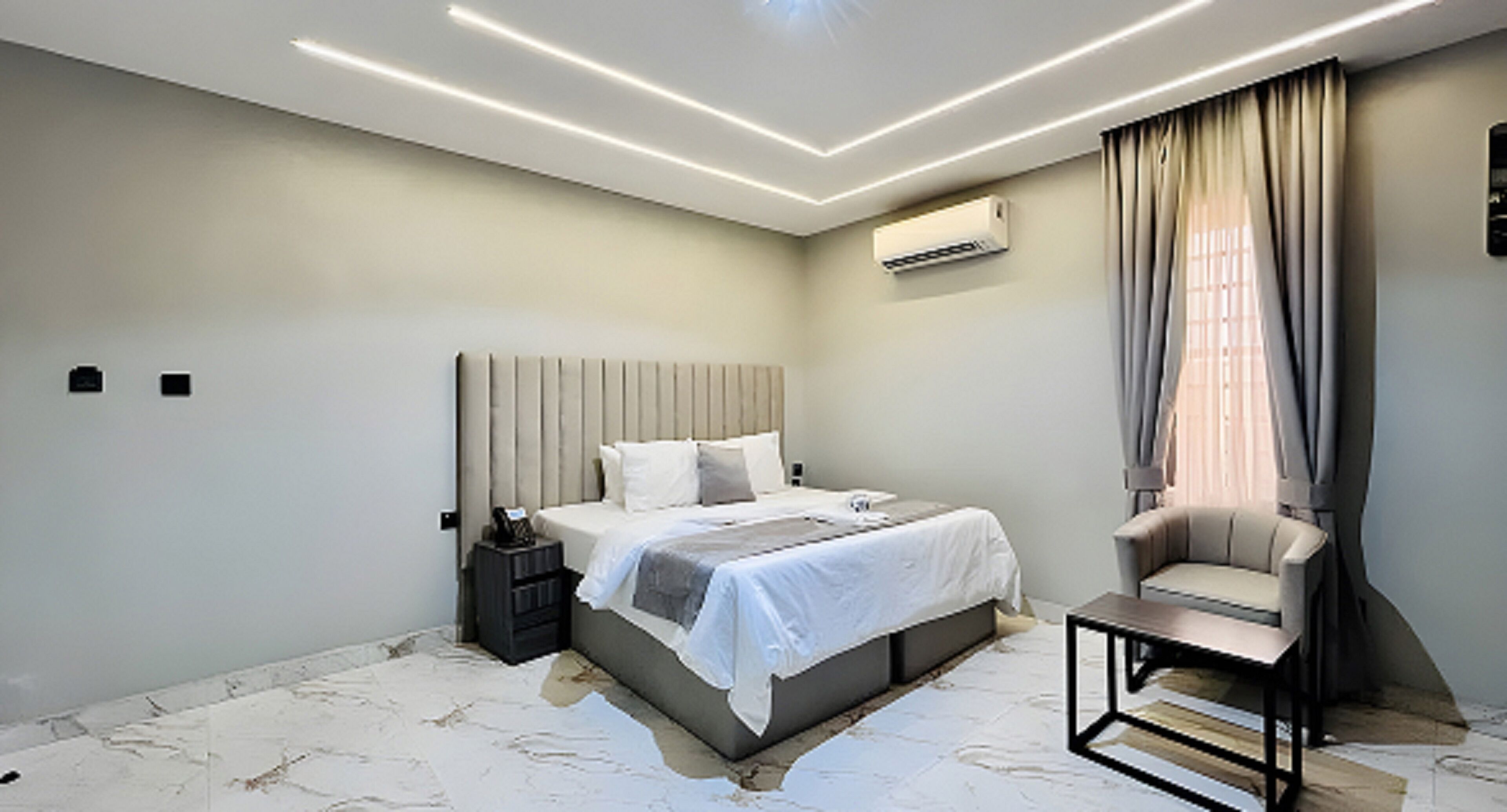 A modern bedroom with a bed, bedside table, chair, and an air conditioning unit.
