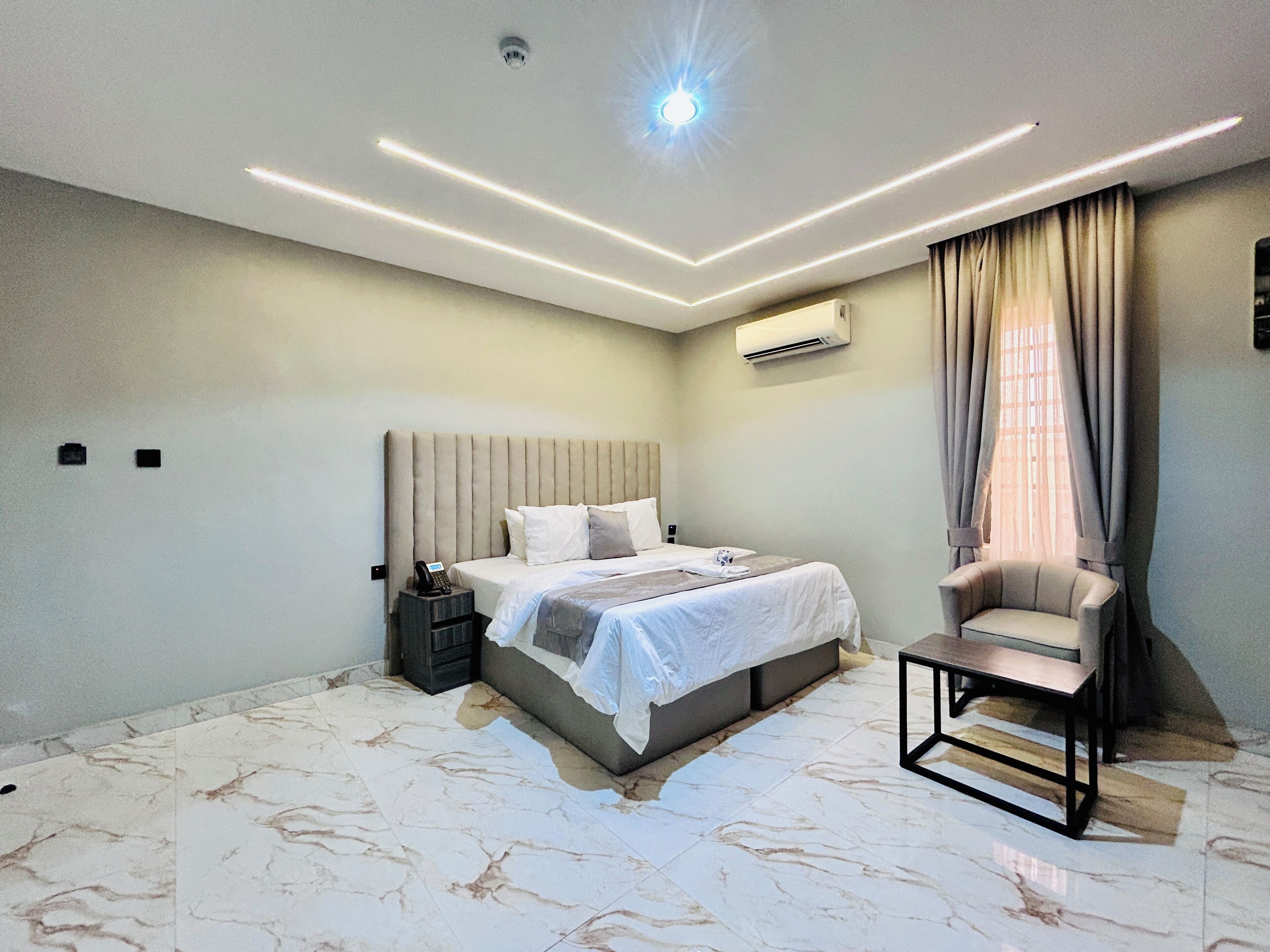 A modern bedroom with a bed, bedside table, chair, and an air conditioning unit.