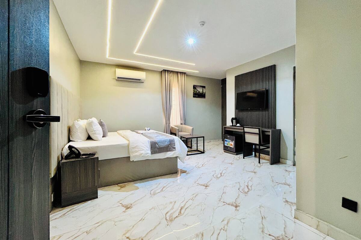 A modern hotel room with a bed, desk, chair, TV, air conditioning unit, and marble flooring.