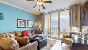 A living room with a sectional sofa, a coffee table, a ceiling fan, and a large sliding glass door leading to a balcony. - 4th-Floor Azure Condo with Free WiFi, Beach Service, Gulf View, Shared Pool,Hot Tub (Fort Walton Beach)