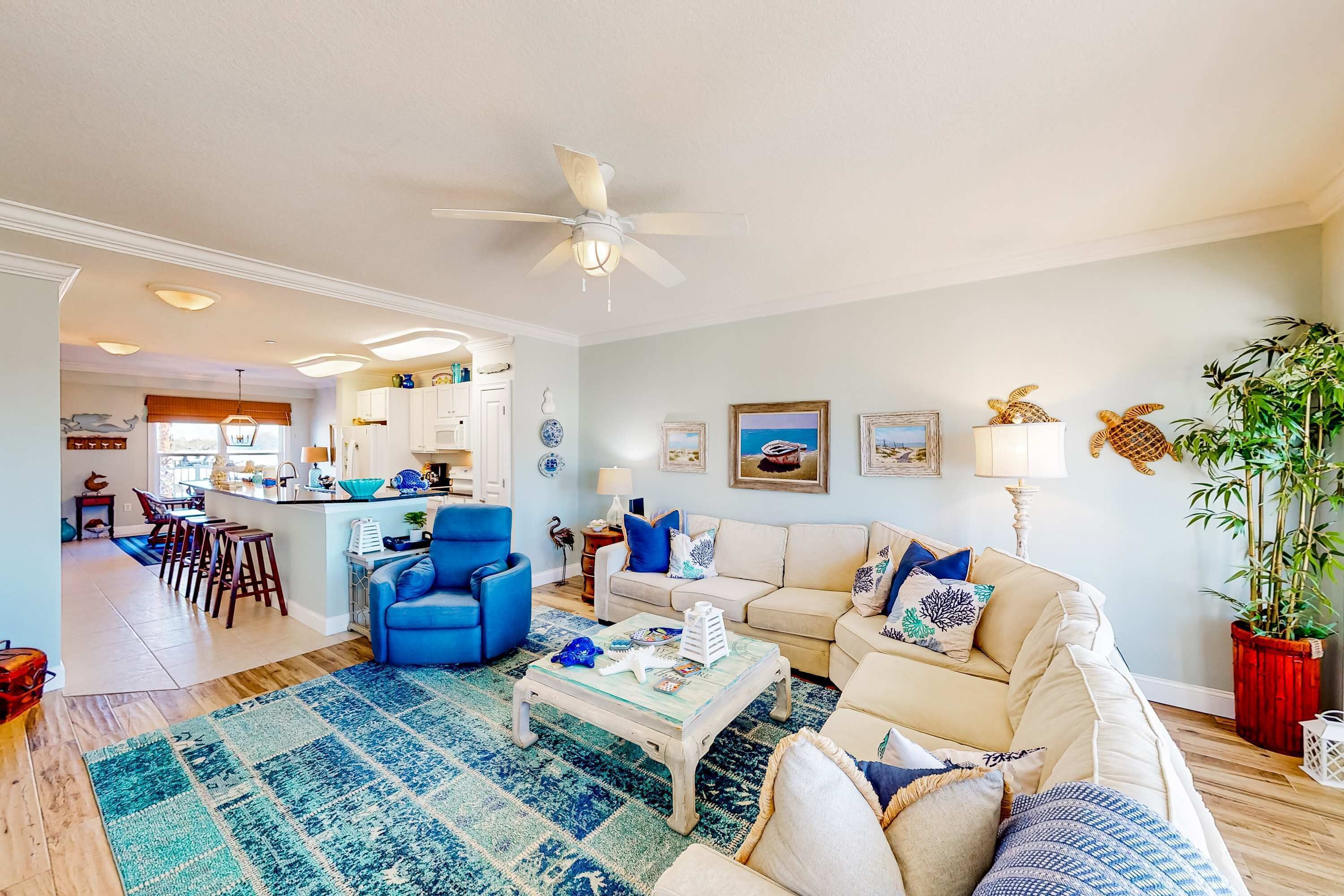 A spacious living room with a large coffee table, a blue armchair, and a beige sofa. There is a kitchen in the background with bar stools.