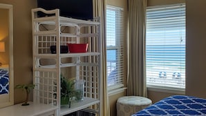 Ett hotellrum med en tv på ett vitt stativ, en säng med ett blått, mönstrat överkast, en liten, rund pall, en vit hylla med dekorativa föremål och fönster med persienner.