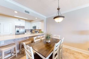 A modern kitchen with white cabinetry, a central island, and a dining area with a wooden table and white chairs. - Gulf Gem with Shared Pool, Hot Tub, Fast WiFi, Fantastic Views & Private Balcony (Fort Walton Beach)