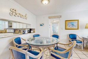 A dining area with a glass-top table surrounded by wicker chairs with blue cushions, a kitchen in the background, and a 'Beach' sign on the wall. - Spacious home with balconies, shared pool and beach access (Fort Walton Beach)