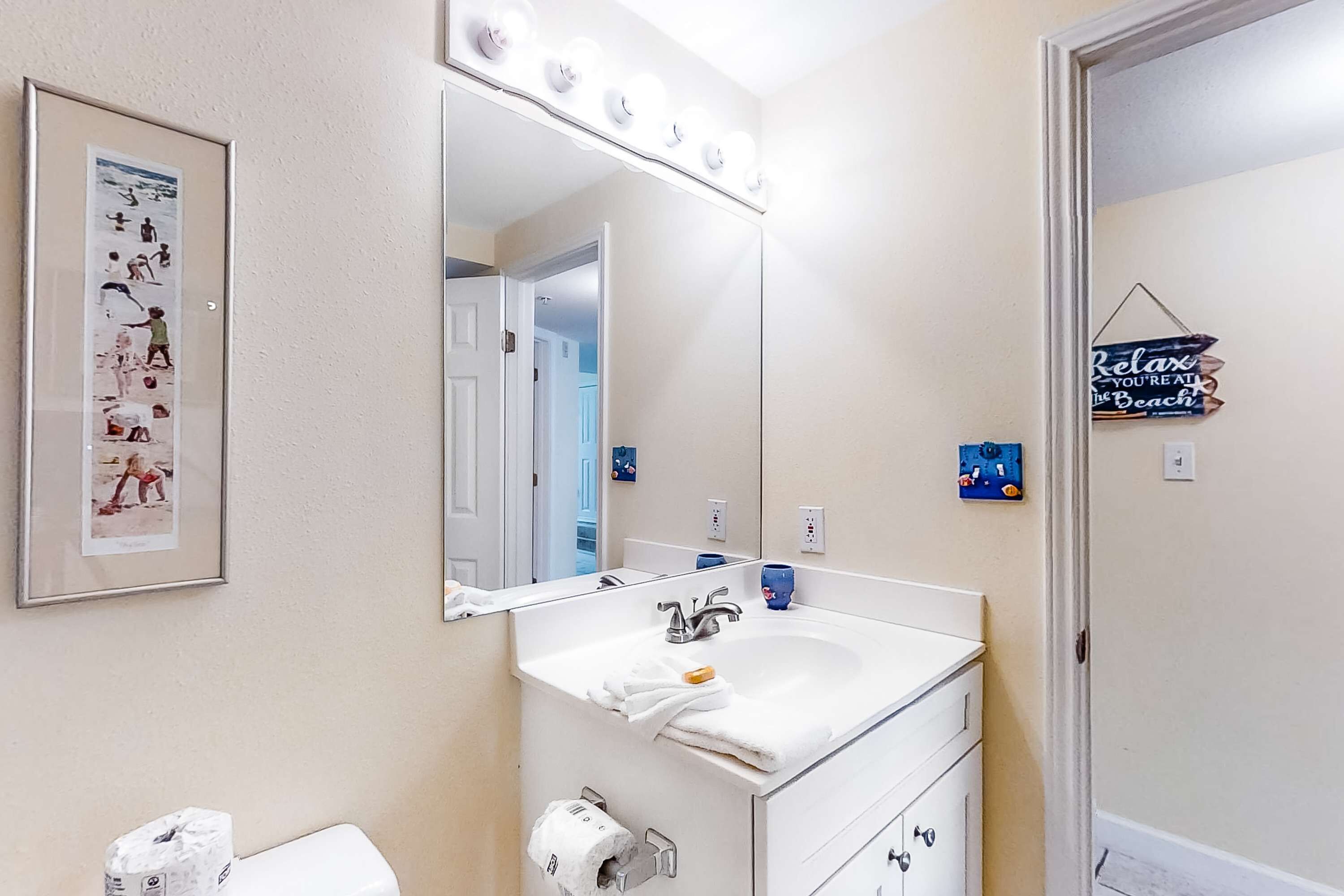 A bathroom with a white sink, mirror, and vanity. There is a framed picture on the wall and a sign that says "Relax, You're on the Beach".