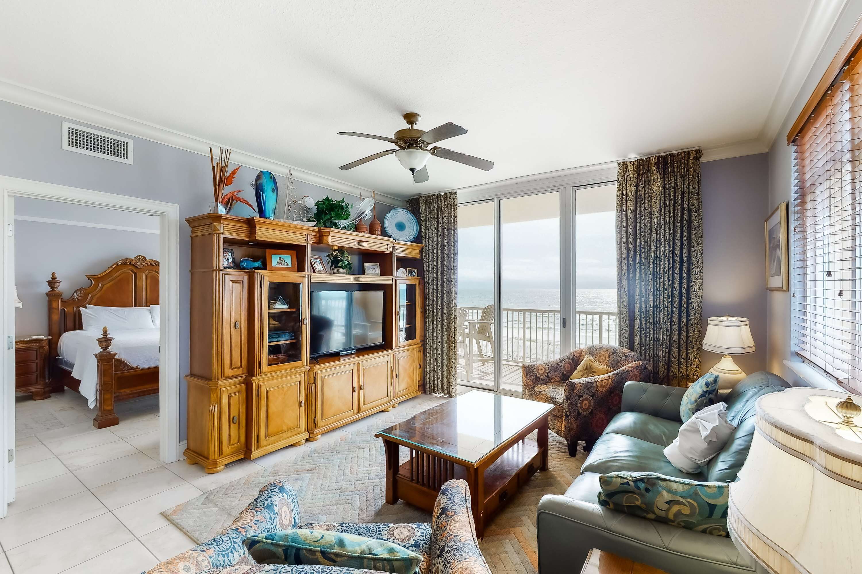A spacious living room with a large wooden cabinet, a television, a ceiling fan, and a view of the outdoors through large windows.