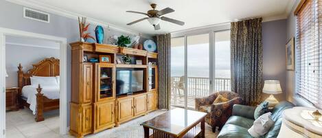 A spacious living room with a large wooden cabinet, a television, a ceiling fan, and a view of the outdoors through large windows.