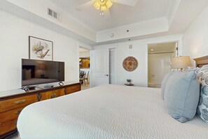A bedroom with a large bed, a TV on a dresser, a ceiling fan, and a door leading to another room. - Beachview Condo with full kitchen, shared pool and beach access (Fort Walton Beach)