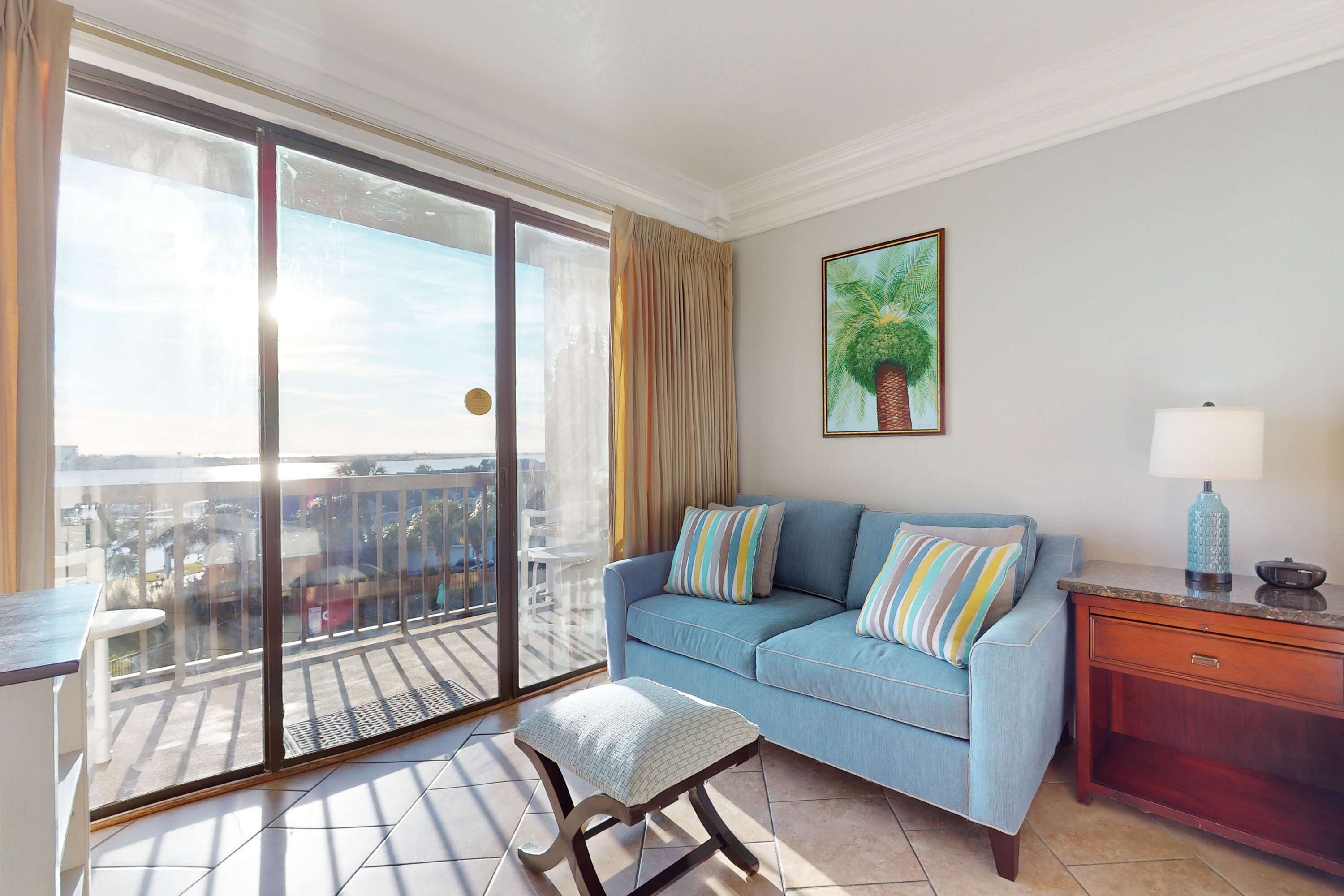 En balkon med skydedør i glas, en blå sofa med stribede puder, et lille bord og en lampe på et træsidebord.