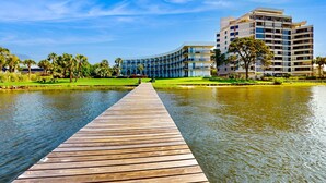 Un muelle de madera que lleva a un edificio moderno de varios pisos, rodeado de vegetación y una masa de agua tranquila.